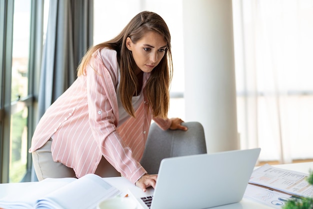 Hermosa mujer de negocios sonriente leyendo algo en una laptop mientras se apoya en la mesa de la oficina