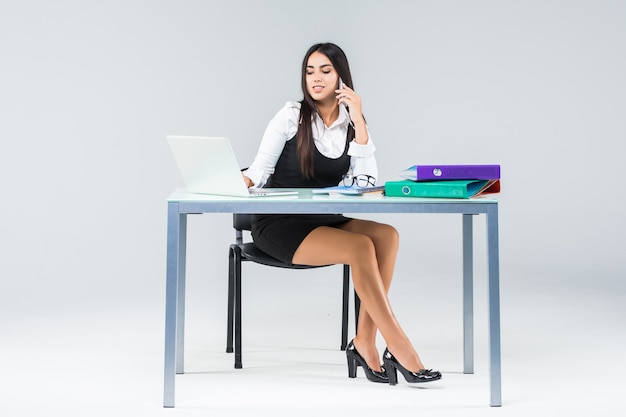 Hermosa mujer de negocios sonriente se sienta desde la mesa y trabaja en la computadora portátil en camisa blanca aislada en blanco.