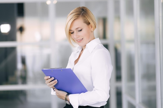 Hermosa mujer de negocios sonriente sosteniendo una carpeta abierta con documentos leyendo mientras está en el cargo