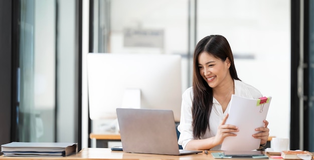 Una hermosa mujer de negocios sonriente usando una computadora portátil y una computadora mientras hace algún papeleo en la oficina