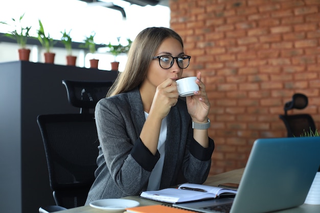 Hermosa mujer de negocios sostiene la taza de café y sonríe mientras está sentada en su lugar de trabajo.