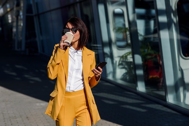 Hermosa mujer de negocios en traje amarillo y gafas de sol caminando cerca del centro de negocios, bebe café y utiliza smartphone. Concepto de negocio, comunicación, gerente y oficinista.