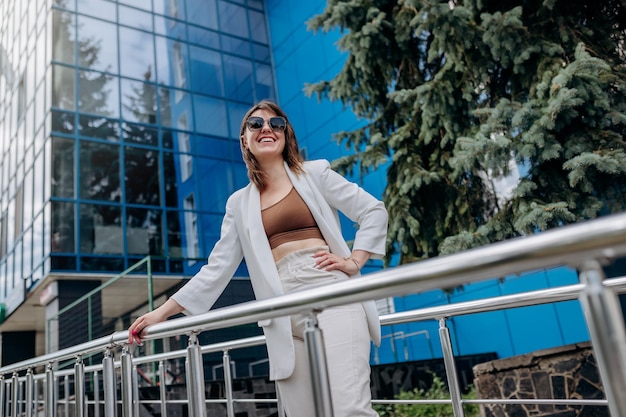 Hermosa mujer de negocios con traje blanco y gafas de sol durante el descanso de pie cerca del moderno edificio de oficinas