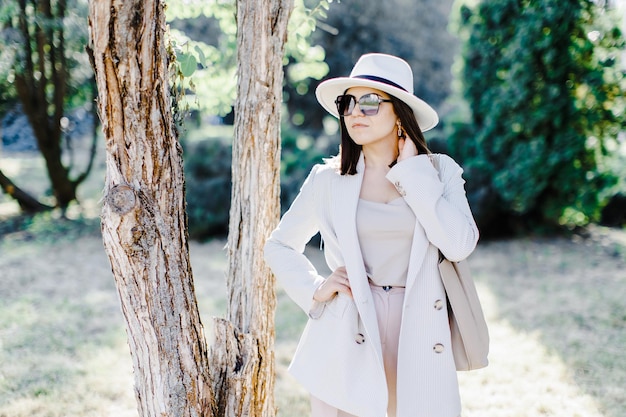 Hermosa mujer de negocios con traje elegante y sombrero de pie cerca de un árbol en el parque