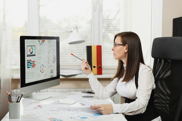 La hermosa mujer de negocios en traje y gafas trabajando en equipo con documentos en la oficina de luz, mirando el monitor