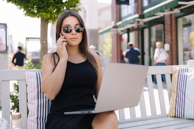 Hermosa mujer de negocios usando teléfono y computadora portátil al mismo tiempo mujer sentada al aire libre en el banco