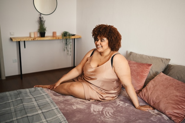 Hermosa mujer negra africana con curvas de talla grande cabello afro acostado en la cama en un vestido rosa de seda en polvo acogedor