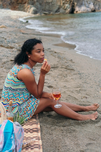 Una hermosa mujer negra bebe vino en la playa, mirando al horizonte.