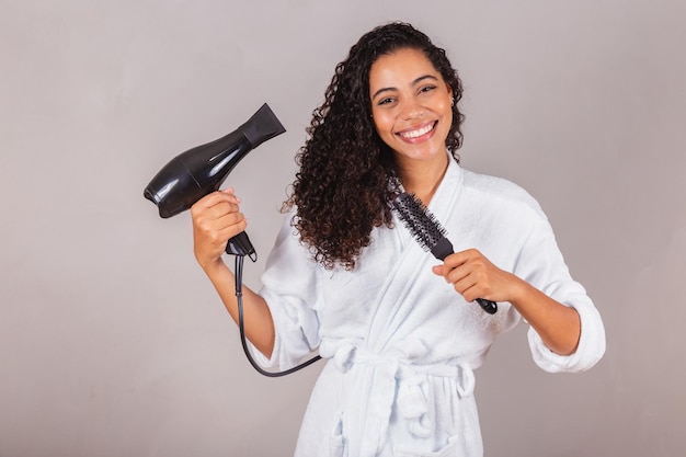 Hermosa mujer negra brasileña con albornoz y toalla secando el cabello con secador y cepillo spa centro de belleza cuidado del cabello