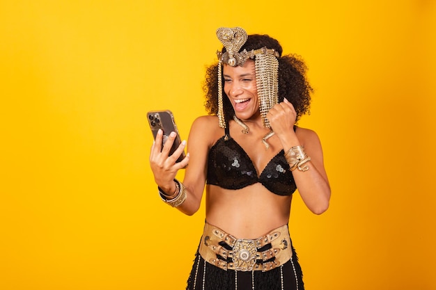 Hermosa mujer negra brasileña con ropa de carnaval de Cleopatra celebrando con smartphone