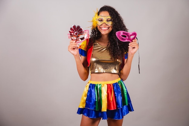 Foto hermosa mujer negra brasileña con ropa de frevo carnaval usando una máscara sosteniendo máscaras de carnaval invitando a la gente a la fiesta