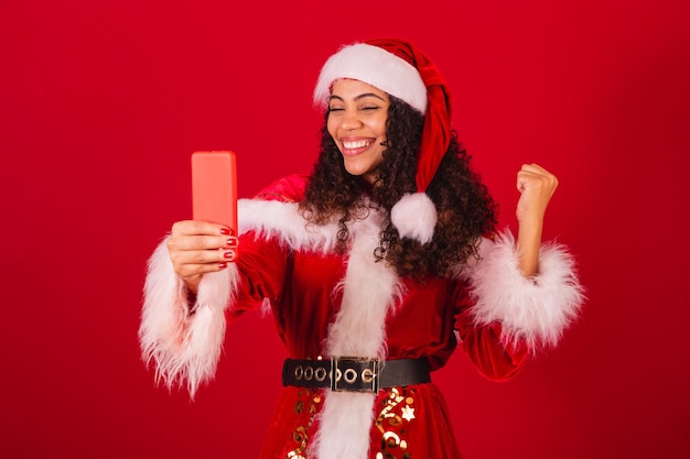 Hermosa mujer negra brasileña vestida como ropa navideña de santa claus celebrando con algo en el teléfono inteligente