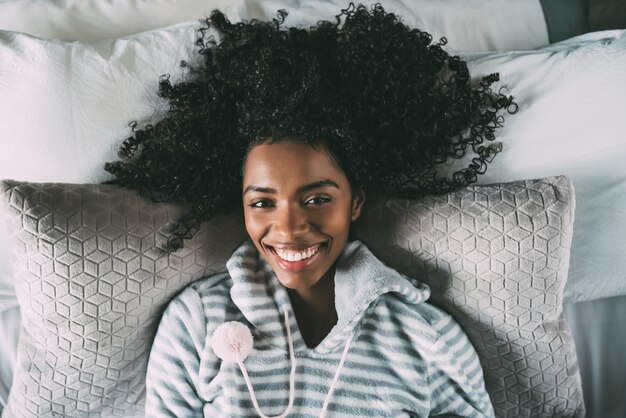 Hermosa mujer negra con cabello rizado sonriendo y acostado en la cama mirando a la cámara