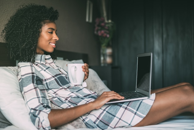 Hermosa mujer negra en cama con laptop y taza de café