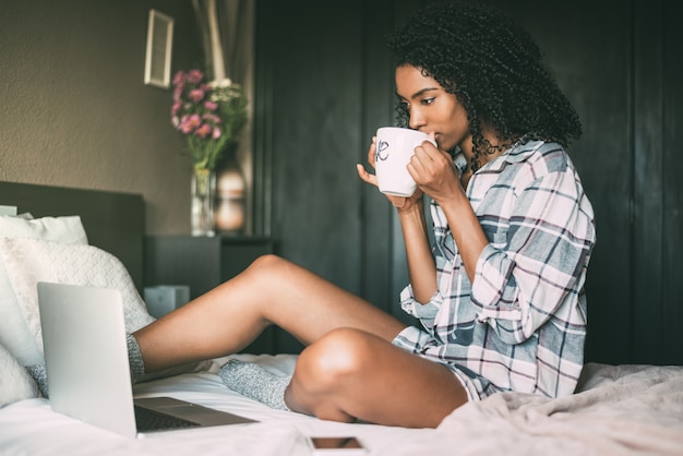 Hermosa mujer negra en cama con laptop y taza de café