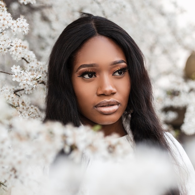 Hermosa mujer negra en colores blancos en la naturaleza. Linda chica encantadora cerca de un árbol en flor. Estilo de vida de belleza natural