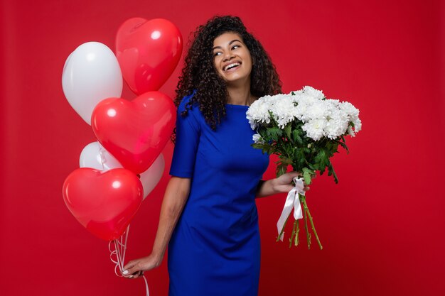 Hermosa mujer negra con globos en forma de corazón y ramo de flores blancas en la pared roja