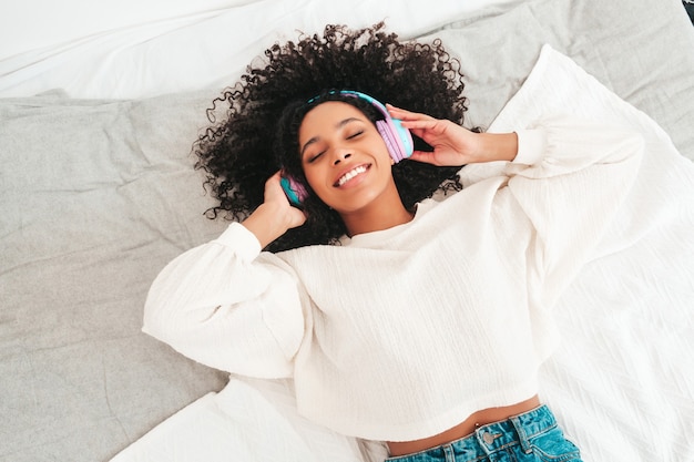 Hermosa mujer negra con peinado afro rizos. Modelo sonriente en suéter y jeans. mujer sin preocupaciones escuchando música en auriculares inalámbricos por la mañana