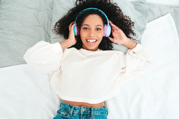 Hermosa mujer negra con peinado afro rizos. Modelo sonriente en suéter y jeans. mujer sin preocupaciones escuchando música en auriculares inalámbricos por la mañana