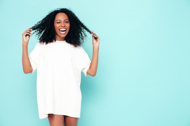 Hermosa mujer negra con peinado de rizos afro Modelo sonriente en ropa de camiseta larga Mujer sexy despreocupada posando junto a la pared azul en el estudio Bronceada y alegre Guiño