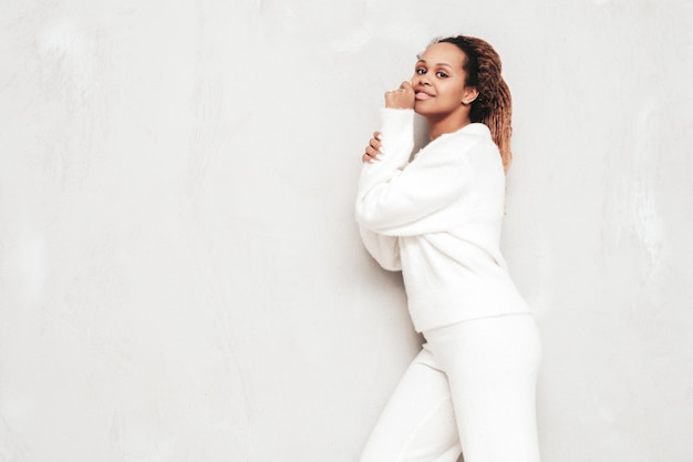 Hermosa mujer negra con peinado de rizos afro Modelo sonriente vestida con traje blanco de verano Sexy mujer despreocupada posando junto a la pared gris en el estudio Bronceada y alegre Sorprendida y sorprendida