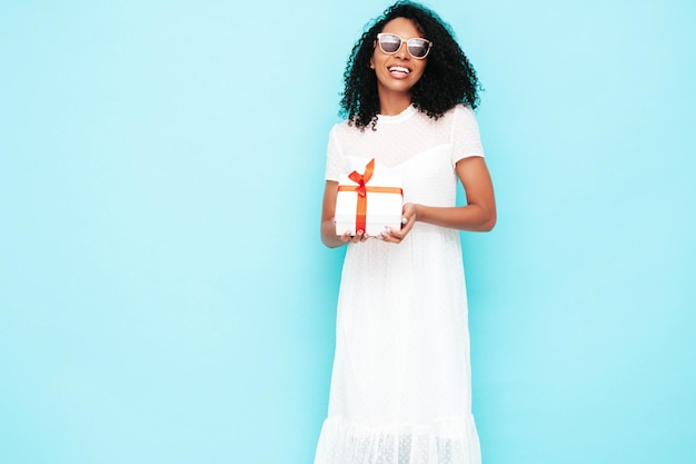 Hermosa mujer negra con peinado de rizos afro Modelo sonriente vestida con vestido blanco de verano Mujer sexy despreocupada posando cerca de la pared azul en el estudio Bronceada y alegre Caja de regalo de sujeción aislada