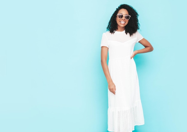 Hermosa mujer negra con peinado de rizos afro Modelo sonriente vestida con vestido blanco de verano Mujer sexy despreocupada posando junto a la pared azul en el estudio Bronceada y alegre con gafas de sol Aislado