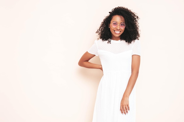 Hermosa mujer negra con peinado de rizos afro Modelo sonriente vestida con vestido blanco de verano Mujer sexy despreocupada posando junto a la pared en el estudio Bronceada y alegre Aislado