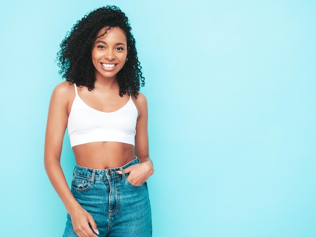 Hermosa mujer negra con peinado de rizos afro Modelo sonriente vestida con vestido blanco de verano Sexy mujer despreocupada posando junto a la pared azul en el estudio Bronceada y alegre Aislado