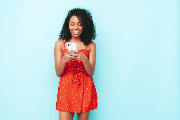Hermosa mujer negra con peinado de rizos afro Modelo sonriente vestida con vestido rojo de verano Sexy mujer despreocupada posando cerca de la pared azul Sosteniendo el teléfono inteligente Mirando la pantalla del teléfono celular Usando aplicaciones