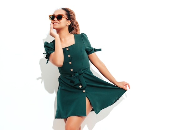 Foto hermosa mujer negra con peinado de rizos afro modelo sonriente vestida con un vestido verde de verano blanco mujer despreocupada sexy posando junto a la pared en el estudio bronceada y alegre aislada en gafas de sol