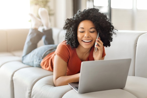 Hermosa mujer negra usando una computadora portátil y hablando por teléfono móvil en casa