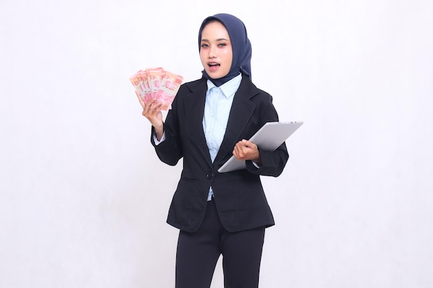 Una hermosa mujer de oficina indonesia con hijab se encuentra conmocionada llevando dinero en rupias y una tableta portátil.