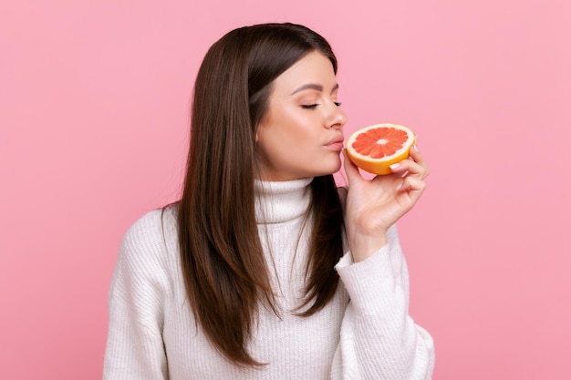 Hermosa mujer oliendo media rodaja de pomelo disfrutando de sabrosas frutas vitamínicas nutrición saludable usando un suéter blanco de estilo casual Foto de estudio interior aislada en fondo rosa