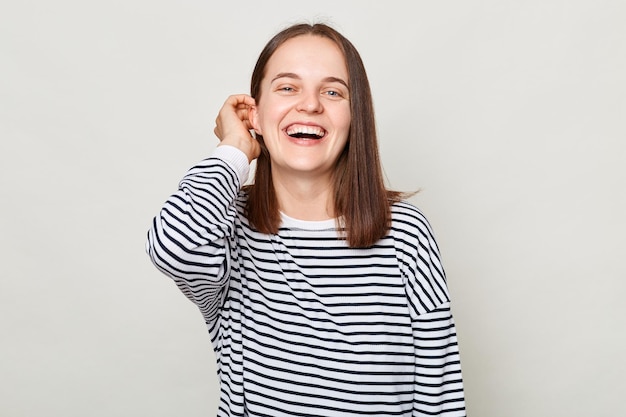 Hermosa mujer optimista con cabello castaño y pantalones a rayas que se encuentra aislada sobre un fondo gris y está de buen humor coqueteando con la mano en el cabello