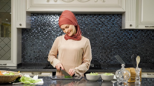 Hermosa mujer oriental en hijab cocina el almuerzo cortando verduras frescas sonriendo en la cocina de la casa closeup