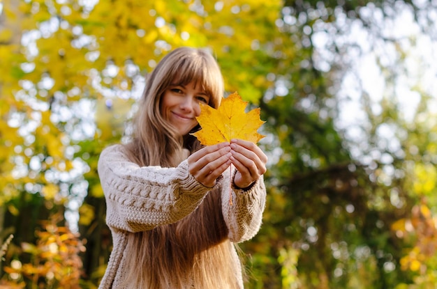 Hermosa mujer de otoño de pie cerca de coloridas hojas de otoño Bastante feliz modelo alegre mirando a la cámara Mujer joven con hoja de otoño en la mano relajación de humor de otoño caminando en la naturaleza