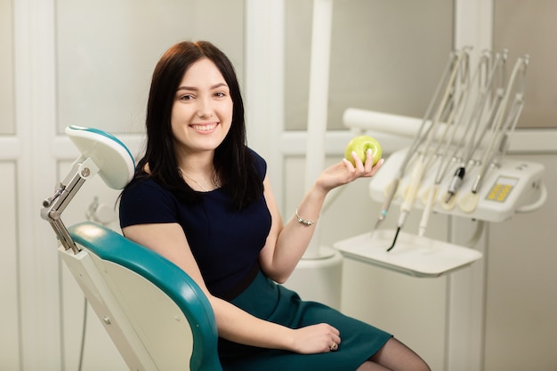 Hermosa mujer paciente sentado en un sillón dental equipo de fondo y sostiene una manzana