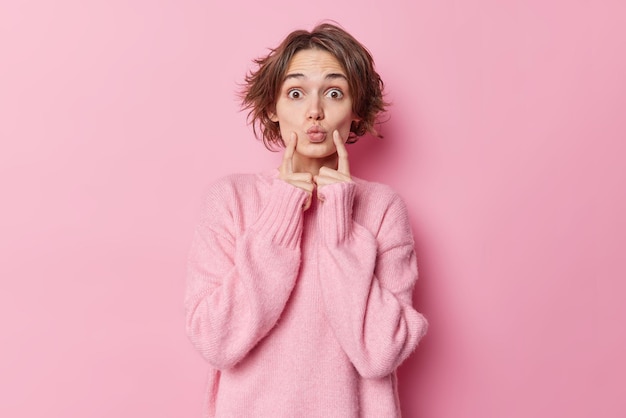 Hermosa mujer con peinado bob mantiene los labios redondeados los dedos índices en las mejillas tiene un aspecto romántico que espera un beso lleva un jersey de cachemir cálido aislado sobre fondo rosa. Concepto de expresiones faciales