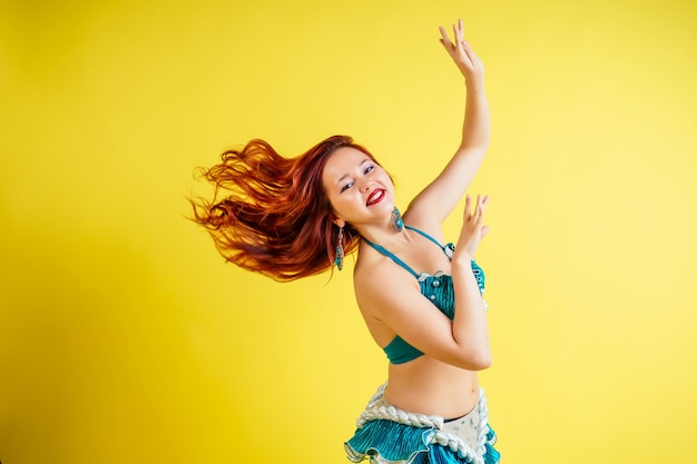 Foto hermosa mujer pelirroja bailando danza del vientre oriental en traje azul sobre fondo amarillo en estudio.