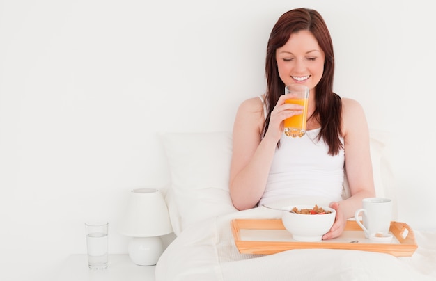 Hermosa mujer pelirroja bebiendo un vaso de jugo de naranja mientras está sentado en su cama