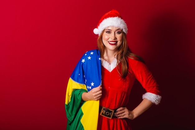 Hermosa mujer pelirroja brasileña vestida con ropa de Navidad Santa Claus sosteniendo la bandera de Brasil