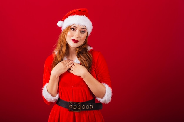 Hermosa mujer pelirroja brasileña vestida con ropa navideña Papá Noel manos en el pecho gratitud