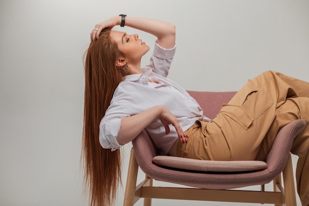 Hermosa mujer pelirroja fresca de moda con cabello rojo natural en ropa elegante de moda sentada y posando en una silla sobre un fondo blanco