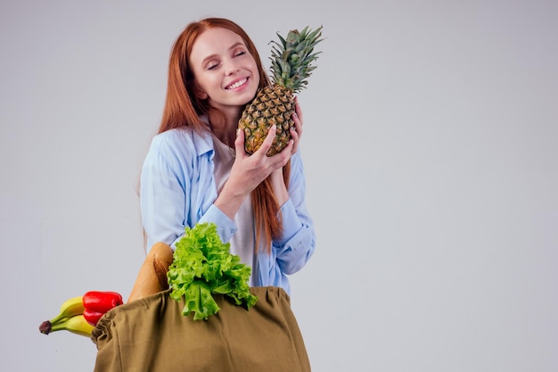 Hermosa mujer pelirroja de jengibre sosteniendo una bolsa de compras de algodón ecológico con frutas frescas, vegetales en el fondo del estudio wgite.