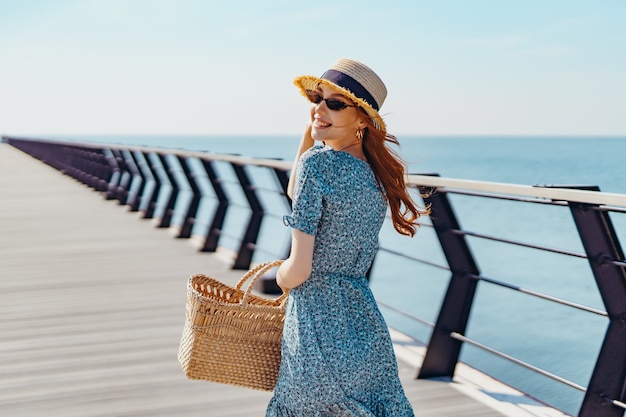 Hermosa mujer pelirroja posando en un día soleado camina por el muelle cerca del mar niña vistiendo fashionab ...