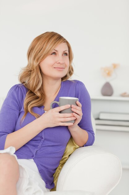 Hermosa mujer pelirroja sosteniendo una taza de café y posando mientras está sentado en un sofá