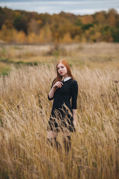 Hermosa mujer pelirroja en un vestido negro camina en un campo de otoño.