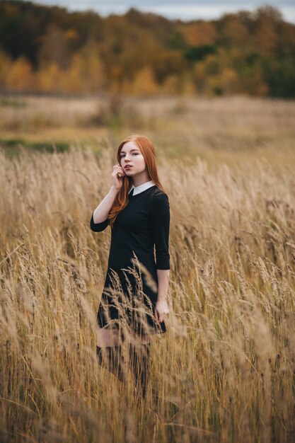 Hermosa mujer pelirroja en un vestido negro camina en un campo de otoño.