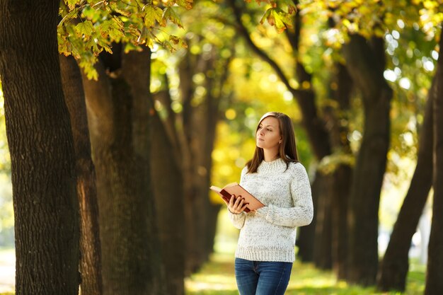 La hermosa mujer de pelo castaño sonriente feliz en suéter blanco de pie con un libro rojo en el parque de la ciudad de otoño en un día cálido. Otoño de hojas doradas. Concepto de lectura.